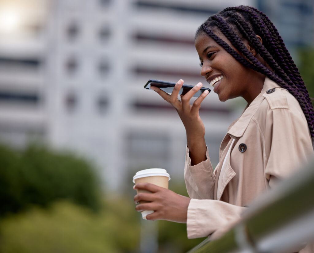 A smiling woman using voice search on her smartphone while holding a coffee cup, representing 2025 marketing trends in AI and digital engagement.